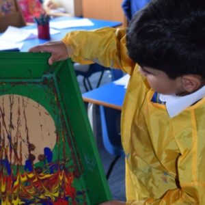 young boy adding lots of different colours of paint to a green container